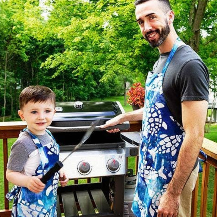 Son Wearing a Kid's Apron with Playful White Turtles and Fish Swimming on an Ocean-Blue Background Helping Dad BBQ | Global Mamas® | Tiny Tastemakers @ Spice Craze®