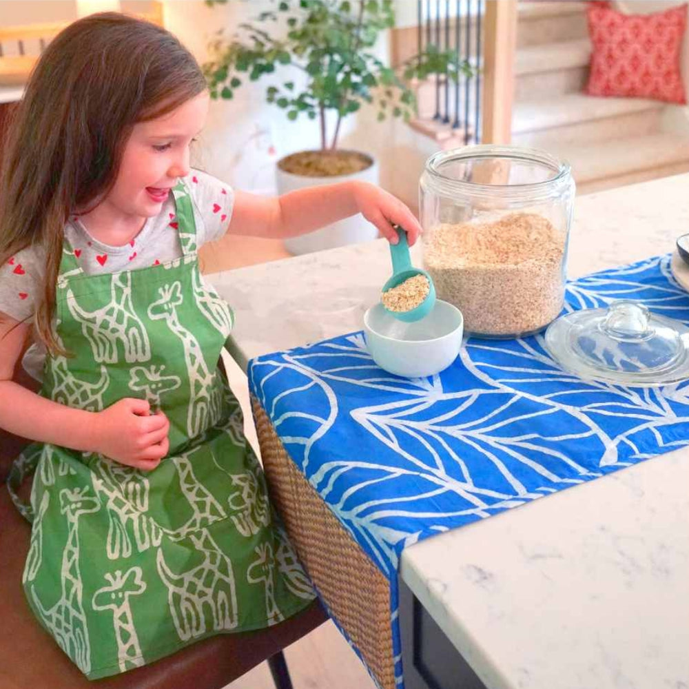 Child Measuring Oatmeal in a Kid's Apron with Charming White Giraffes on a Bright Olive-Green Background | Global Mamas® | Tiny Tastemakers @ Spice Craze®